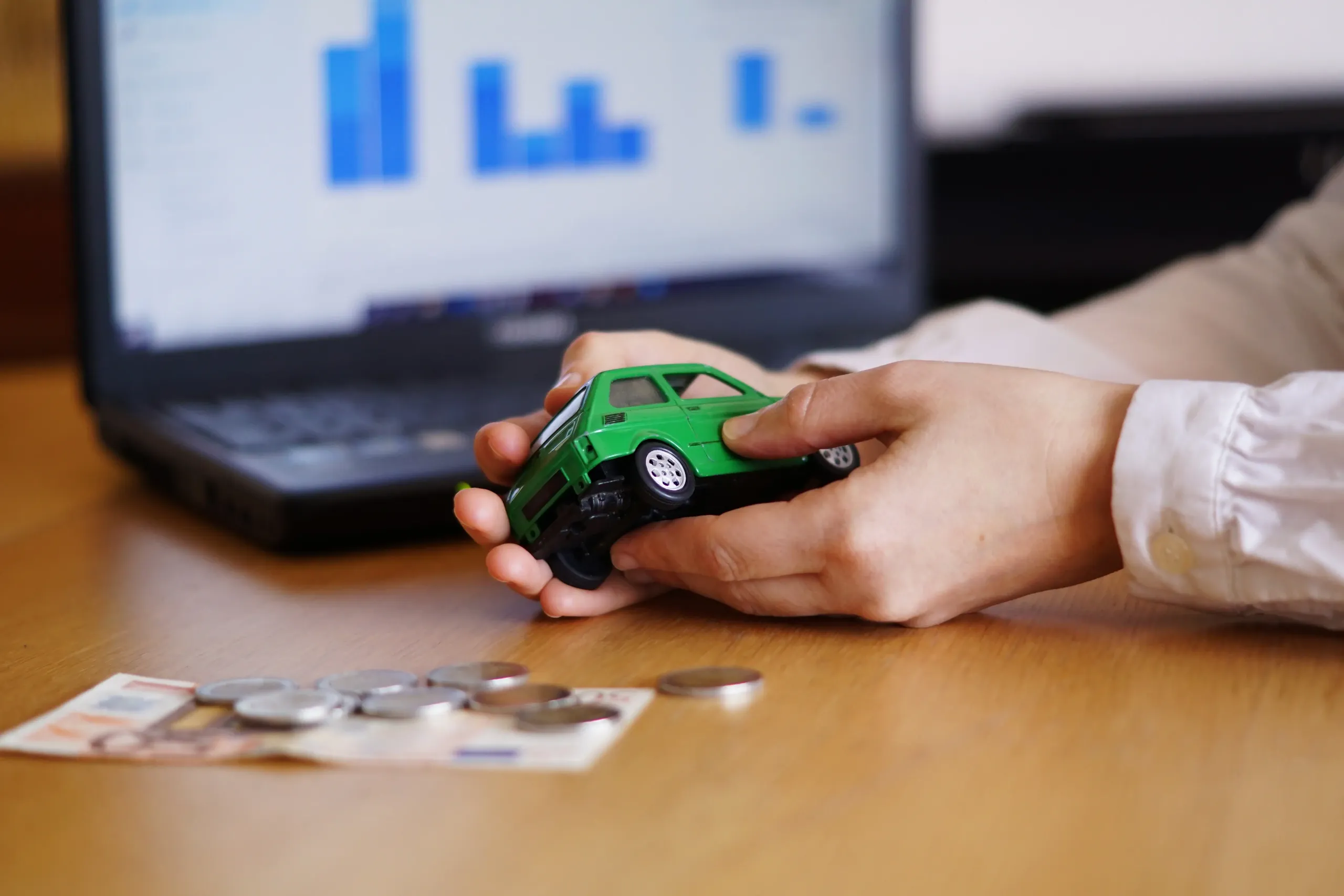 homem com carro de brincar nas mãos, dinheiro sobre a mesa e computador com gráficos atrás